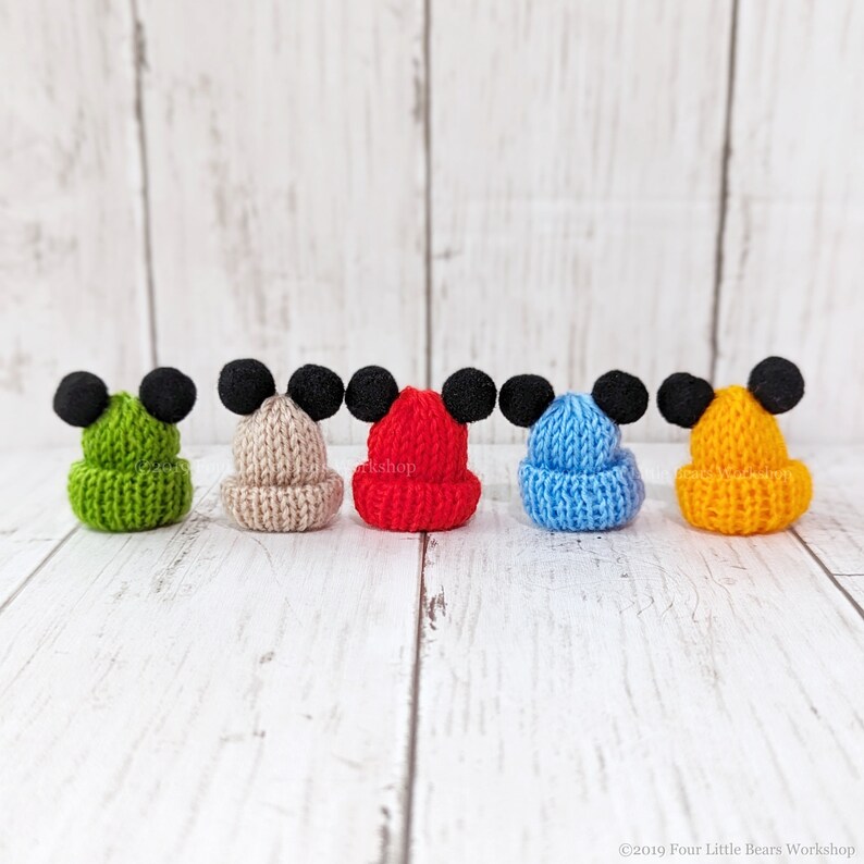 Pu&ograve; includere: Cinque cappellini in miniatura di maglia con pompon neri in cima, che ricordano le orecchie di Topolino. I cappellini sono di diversi colori: verde, beige, rosso, blu e giallo.