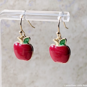 May include: A pair of gold dangle earrings with red apple charms. The apples have green leaves and are outlined in gold.