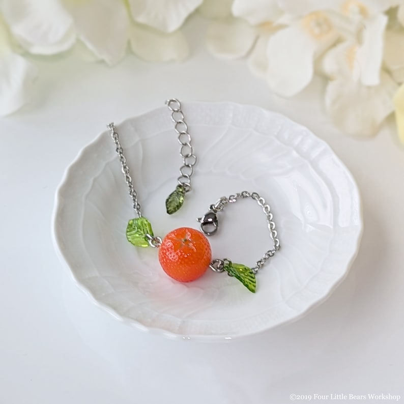 May include: A silver chain bracelet with a small orange fruit charm and green leaf charms. The bracelet is on a white plate.