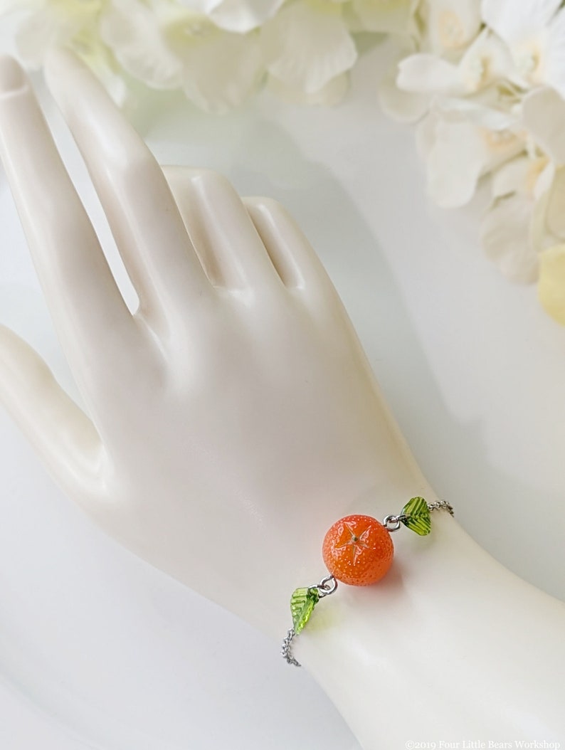 May include: A silver chain bracelet with a single orange charm and green leaf charms. The bracelet is on a white mannequin wrist.