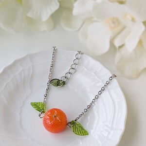 May include: A silver chain necklace with a small orange charm and green leaf accents. The necklace is displayed on a white plate.