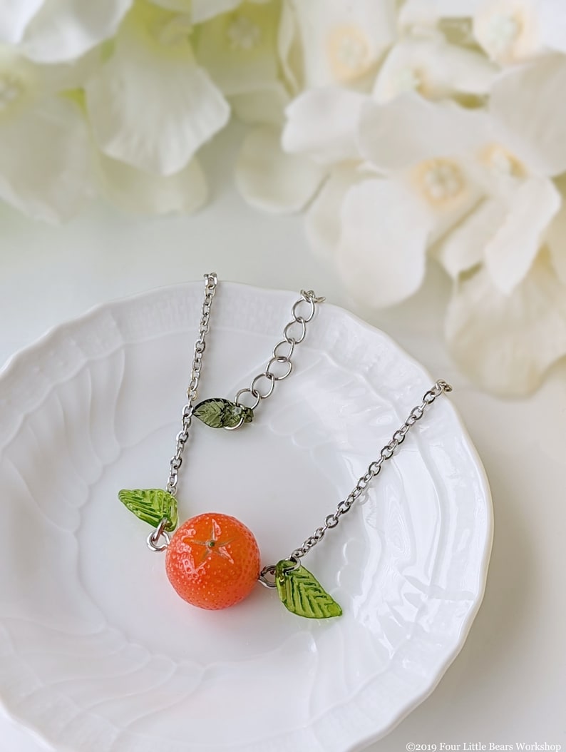 May include: A silver chain necklace with a small orange charm and green leaf accents. The necklace is displayed on a white plate.