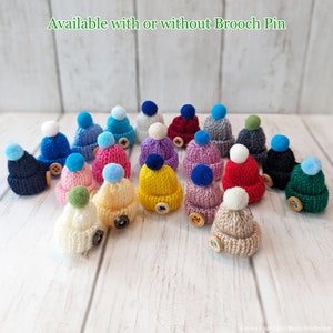 May include: A collection of 18 miniature knitted hats in various colors, including pink, blue, yellow, green, and red. Each hat has a small pom-pom on top and a button on the side. The hats are available with or without a brooch pin.
