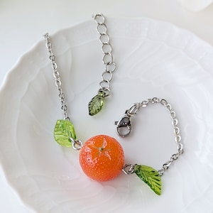 May include: A silver chain bracelet with a small orange charm and green leaf charms. The orange charm is detailed with a star-shaped design.
