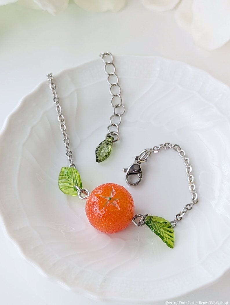 May include: A silver chain bracelet with a small orange charm and green leaf charms. The orange charm is detailed with a star-shaped design.