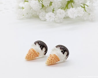May include: A pair of small, white and brown ice cream cone earrings. The ice cream is white with a brown chocolate topping. The cone is a light brown color.