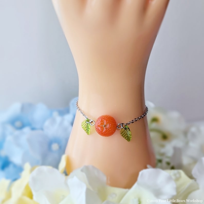 May include: A silver chain bracelet with a small orange slice charm and two green leaf charms.