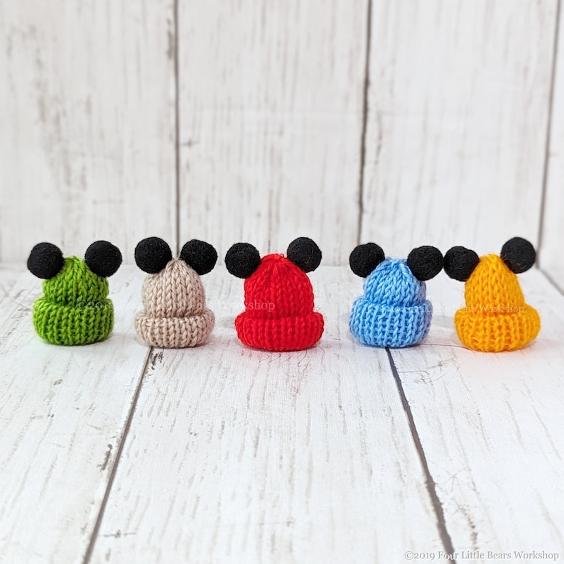 Pu&ograve; includere: Cinque cappellini in maglia in miniatura con pompon neri in cima. I cappellini sono verde, beige, rosso, blu e giallo.