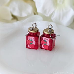 May include: A pair of red strawberry jam jar earrings. Each miniature jar has a gold lid and a label with the text "JAM STRAWBERRY" and a strawberry graphic. The earrings are attached to silver-colored hooks.