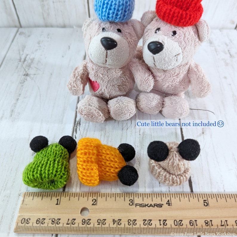 Pu&ograve; includere: Quattro cappellini in miniatura di lana con pon pon neri. I cappellini sono verde, arancione, beige e blu. I cappellini sono disposti su un righello di legno con segni in pollici. Il testo "Cute little bears not included" &egrave; scritto sotto i cappellini.