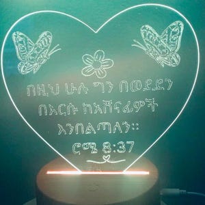 May include: A heart-shaped LED night light with a wooden base. The light features two butterflies, a flower, and the text "ሰው ሁሉ ጌታ በመሆኑ በእርሱ አእምሮ ፍቃድ አገልግሉ:: ሮሜ 8:37".