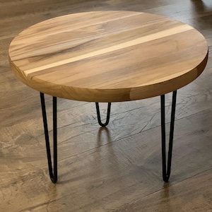 May include: A round wooden side table with a light brown finish. The tabletop is supported by three black hairpin legs. The table is approximately 20 inches in diameter and 18 inches tall.