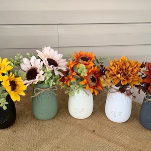May include: Five mason jars painted in different colors, each holding a different type of fall flower arrangement. The jars are arranged in a row on a burlap surface. The flowers include sunflowers, white daisies, orange mums, and red mums.