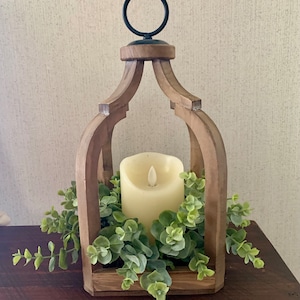 May include: A wooden lantern with a black metal ring on top. The lantern is filled with greenery and a white pillar candle.