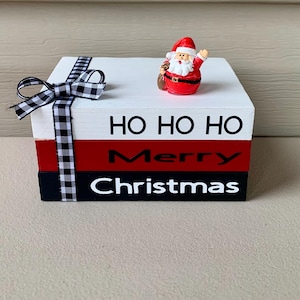 May include: A white, red, and black wooden box with the words "Ho Ho Ho", "Merry", and "Christmas" written on it. A small Santa Claus figurine is on top of the box.
