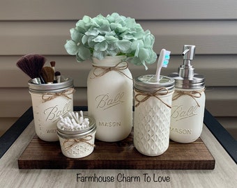 May include: Set of five white painted mason jars with lids, used as bathroom organizers. The jars are decorated with twine and a decorative design. The jars are on a wooden tray. The text "Farmhouse Charm To Love" is on the tray.