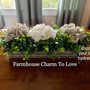 May include: A farmhouse-style centerpiece featuring three hydrangeas in a galvanized metal tray. The hydrangeas are in shades of white and gray, and the tray is surrounded by greenery. The text "Choose your color hydrangea" and "Farmhouse Charm To Love" is visible in the image.
