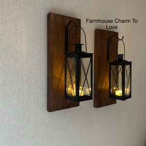 May include: Two black metal lanterns with candle holders are mounted on wooden plaques. The plaques are attached to a white wall. The text "Farmhouse Charm To Love" is written in white on the wall.