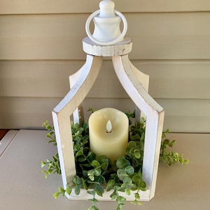 May include: White wooden lantern with a candle inside and greenery around the base. The lantern has a white handle on top.