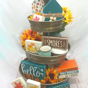 Op de afbeelding: Een metalen dienblad met drie niveaus, gevuld met herfstthema-decoraties. Inclusief houten borden met zinnen als "S'mores" en "Hello Fall", zonnebloemen en een klein houten huis. Een banner met het woord "FALL" staat onderaan.