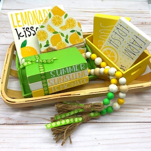 Puede incluir: Un arreglo decorativo con temática de limón, que incluye bloques y cuentas de madera. Los bloques están pintados de amarillo y verde, con frases como "Summer Sunshine Lemonade". También se incluye una guirnalda de cuentas de madera y una borla decorativa.