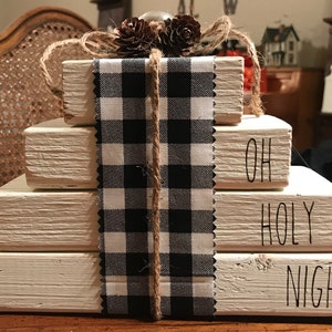 May include: A decorative wooden block set with the words "OH HOLY NIGHT" painted on the sides. The blocks are stacked and wrapped with a black and white checkered ribbon and twine. Pinecones and a silver ornament adorn the top.