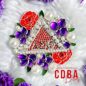 May include: A silver and red sorority pin with a triangle design and the letters "ΔΣΘ" surrounded by pearls and flowers. The flowers are red and purple with green leaves. The pin is on a white fluffy surface.