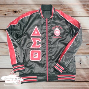 May include: Black satin bomber jacket with red and white stripes on the sleeves and bottom hem. The jacket has a zipper closure and features a large embroidered Greek letter design on the front. The text "Delta Sigma Theta Sorority, Inc." is also embroidered on the jacket.