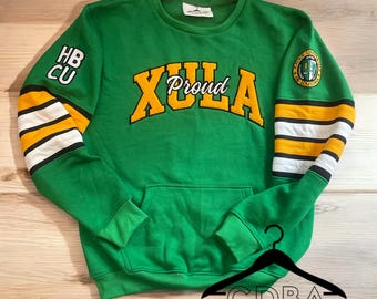 May include: Green sweatshirt with yellow and white striped sleeves. The front of the sweatshirt has the words "XULA Proud" in yellow and black lettering. The left sleeve has the letters "HB CU" in black lettering. The right sleeve has a small embroidered logo with the words "XULA University" in black lettering.