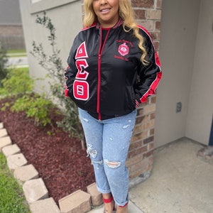 May include: Black satin bomber jacket with red and white trim. The jacket features a large embroidered Greek letter design on the front and a small embroidered logo on the right side. The jacket is zipped up and the model is wearing blue jeans and red heels.