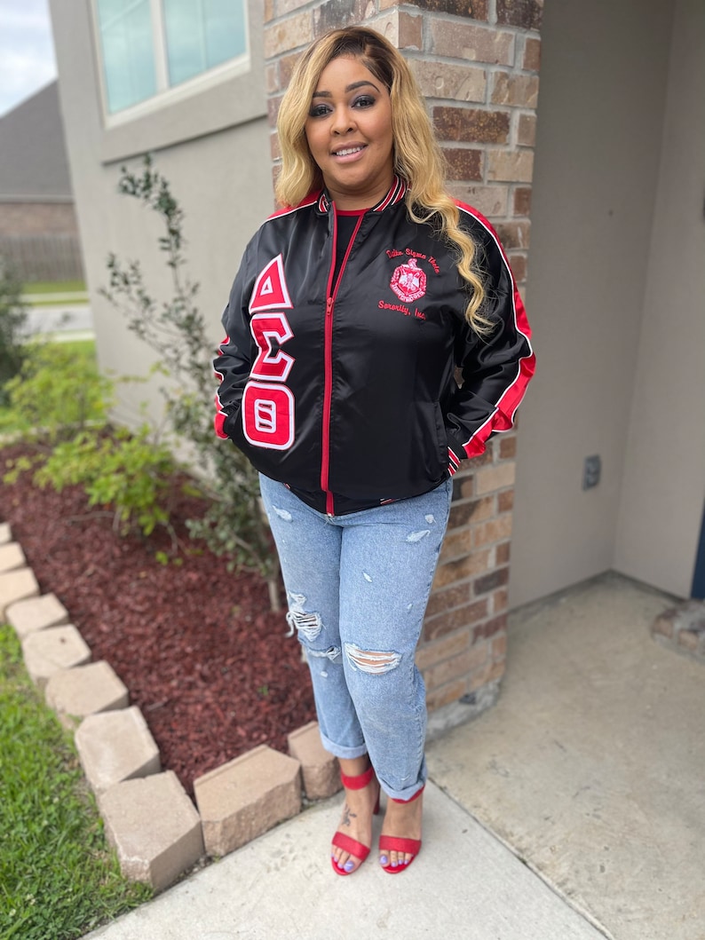 May include: Black satin bomber jacket with red and white trim. The jacket features a large embroidered Greek letter design on the front and a small embroidered logo on the right side. The jacket is zipped up and the model is wearing blue jeans and red heels.