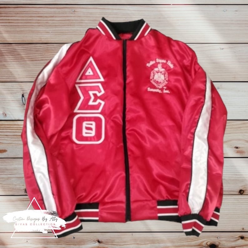 May include: Red satin bomber jacket with white and black striped sleeves and a black zipper. The jacket has a white embroidered Greek letter design on the left sleeve and a white embroidered logo on the right side of the chest. The logo reads "Delta Sigma Theta Sorority, Inc."
