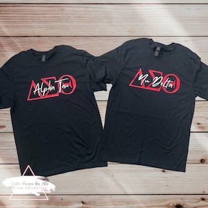 May include: Two black t-shirts with red Greek letters and white text. The left shirt says "Alpha Tau" and the right shirt says "Nu Delta".
