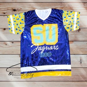 May include: Blue sequined jersey with yellow and blue leopard print sleeves. The jersey has the letters "SU" in blue and yellow with a light blue outline. The text "Jaguars 1880" is written in white below the letters.