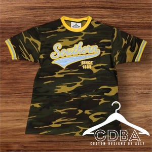 May include: Camouflage pattern t-shirt with yellow collar and sleeve trim. The shirt features the word "Southern" in a script font with a light blue accent, and the text "SINCE 1888". The shirt is displayed on a white hanger.