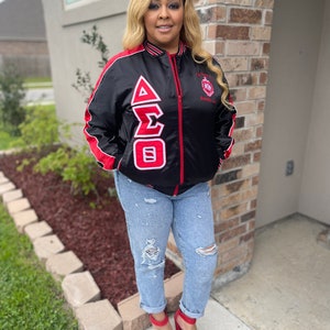May include: A black satin bomber jacket with red and white trim. The jacket features a large embroidered Greek letter design on the left side and a small embroidered crest on the right side. The jacket is worn by a person with long blonde hair and red shoes.
