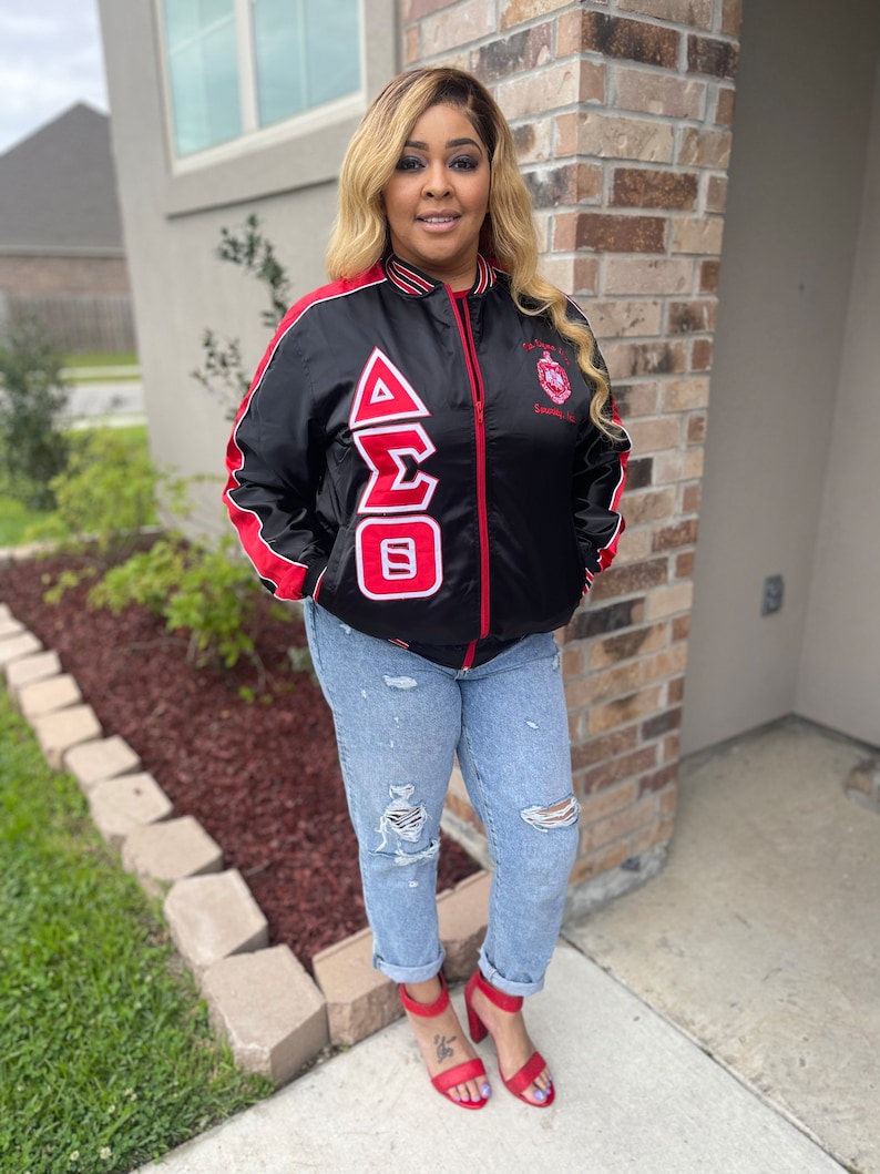 May include: A black satin bomber jacket with red and white trim. The jacket features a large embroidered Greek letter design on the left side and a small embroidered crest on the right side. The jacket is worn by a person with long blonde hair and red shoes.