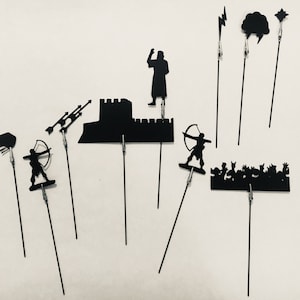 May include: A collection of black silhouette cutouts on wire stems, including a castle, archer, and lightning bolt. The cutouts are held in place with small clips, creating a unique display against a white background.
