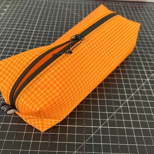 Peut inclure: Une trousse rectangulaire en tissu orange vif avec un motif &agrave; carreaux blancs. La trousse est dot&eacute;e d'une fermeture &eacute;clair noire sur le dessus, &eacute;quip&eacute;e d'une tirette en cordon noir. Une petite &eacute;tiquette en tissu noir avec du texte blanc est fix&eacute;e.