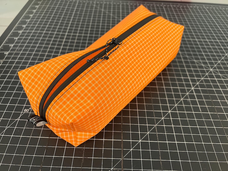 Peut inclure: Une trousse rectangulaire en tissu orange vif avec un motif &agrave; carreaux blancs. La trousse est dot&eacute;e d'une fermeture &eacute;clair noire sur le dessus, &eacute;quip&eacute;e d'une tirette en cordon noir. Une petite &eacute;tiquette en tissu noir avec du texte blanc est fix&eacute;e.