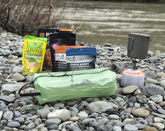 Puede incluir: Un conjunto de alimentos para acampar, que incluyen macarrones con queso, granola y una olla pequeña, están dispuestos sobre un lecho de piedras de río lisas. También se puede ver una pequeña estufa de camping y una bolsa ultraligera verde.