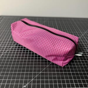 Peut inclure: Une trousse rectangulaire zipp&eacute;e avec un motif &agrave; carreaux rose et blanc. Le sac poss&egrave;de une fermeture &eacute;clair noire sur le dessus et une petite &eacute;tiquette en tissu blanc sur le c&ocirc;t&eacute; avec le texte ULTRALITE imprim&eacute; en lettres noires.