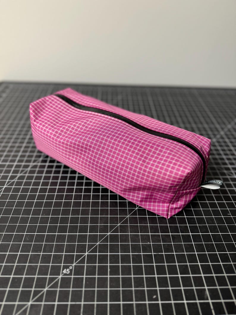 Peut inclure: Une trousse rectangulaire zipp&eacute;e avec un motif &agrave; carreaux rose et blanc. Le sac poss&egrave;de une fermeture &eacute;clair noire sur le dessus et une petite &eacute;tiquette en tissu blanc sur le c&ocirc;t&eacute; avec le texte ULTRALITE imprim&eacute; en lettres noires.