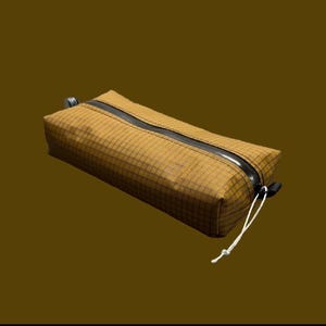 Peut inclure: Une trousse rectangulaire jaune moutarde avec un motif &agrave; carreaux sur tout le tissu. La trousse comprend une fermeture &eacute;clair noire en diagonale sur le dessus et un cordon blanc fin attach&eacute; &agrave; la tirette pour une ouverture facile.