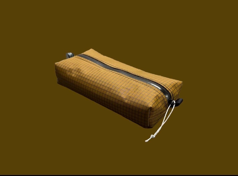 Peut inclure: Une trousse rectangulaire jaune moutarde avec un motif &agrave; carreaux sur tout le tissu. La trousse comprend une fermeture &eacute;clair noire en diagonale sur le dessus et un cordon blanc fin attach&eacute; &agrave; la tirette pour une ouverture facile.
