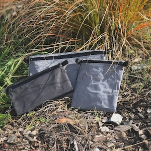 Pode incluir: Três bolsas retangulares de tamanhos e tons de cinza variados. Cada bolsa possui um fecho de correr preto na borda superior. O material tem uma aparência enrugada e texturizada, dispostas em um grupo sobre o solo.