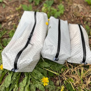 Peut inclure: Trois pochettes de rangement blanches de tailles diff&eacute;rentes, chacune pr&eacute;sentant un motif &agrave; grille noire distinct et une fermeture &eacute;clair noire verticale au centre. Chaque pochette comporte une petite &eacute;tiquette en tissu noir et blanc.
