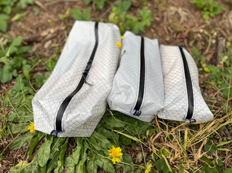 Peut inclure: Trois pochettes de rangement blanches de tailles diff&eacute;rentes, chacune pr&eacute;sentant un motif &agrave; grille noire distinct et une fermeture &eacute;clair noire verticale au centre. Chaque pochette comporte une petite &eacute;tiquette en tissu noir et blanc.