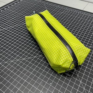 Peut inclure: Une trousse rectangulaire vert citron avec un motif &agrave; carreaux blancs et une fermeture &eacute;clair noire r&eacute;sistante &agrave; l'eau sur le dessus. La trousse poss&egrave;de une petite languette en tissu noir &agrave; une extr&eacute;mit&eacute; et deux tirettes noires avec des cordons.