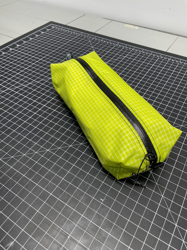 Peut inclure: Une trousse rectangulaire vert citron avec un motif &agrave; carreaux blancs et une fermeture &eacute;clair noire r&eacute;sistante &agrave; l'eau sur le dessus. La trousse poss&egrave;de une petite languette en tissu noir &agrave; une extr&eacute;mit&eacute; et deux tirettes noires avec des cordons.